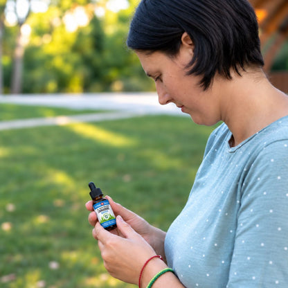 Vrouw in een park die het etiket van een flesje CBD-olie leest, in een lichtblauw shirt met stippen